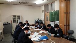 スーツ姿の8人が会議室で資料をチェックしている様子の写真