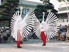 赤いズボンと白い羽を模した鶴の姿をした二人が両手を広げて向かい合っている写真