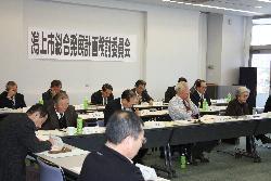 壁に潟上市総合発展計画検討委員会と書かれた紙が貼ってあり、壁の手前に三列に机が並べられ人々が座っている写真