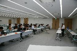 大会議室で会議をしている司会進行の2人と参加している各班長の様子の写真