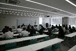 大会議室で会議をしている司会進行の2人と参加している各班長の様子を後方から撮影した写真