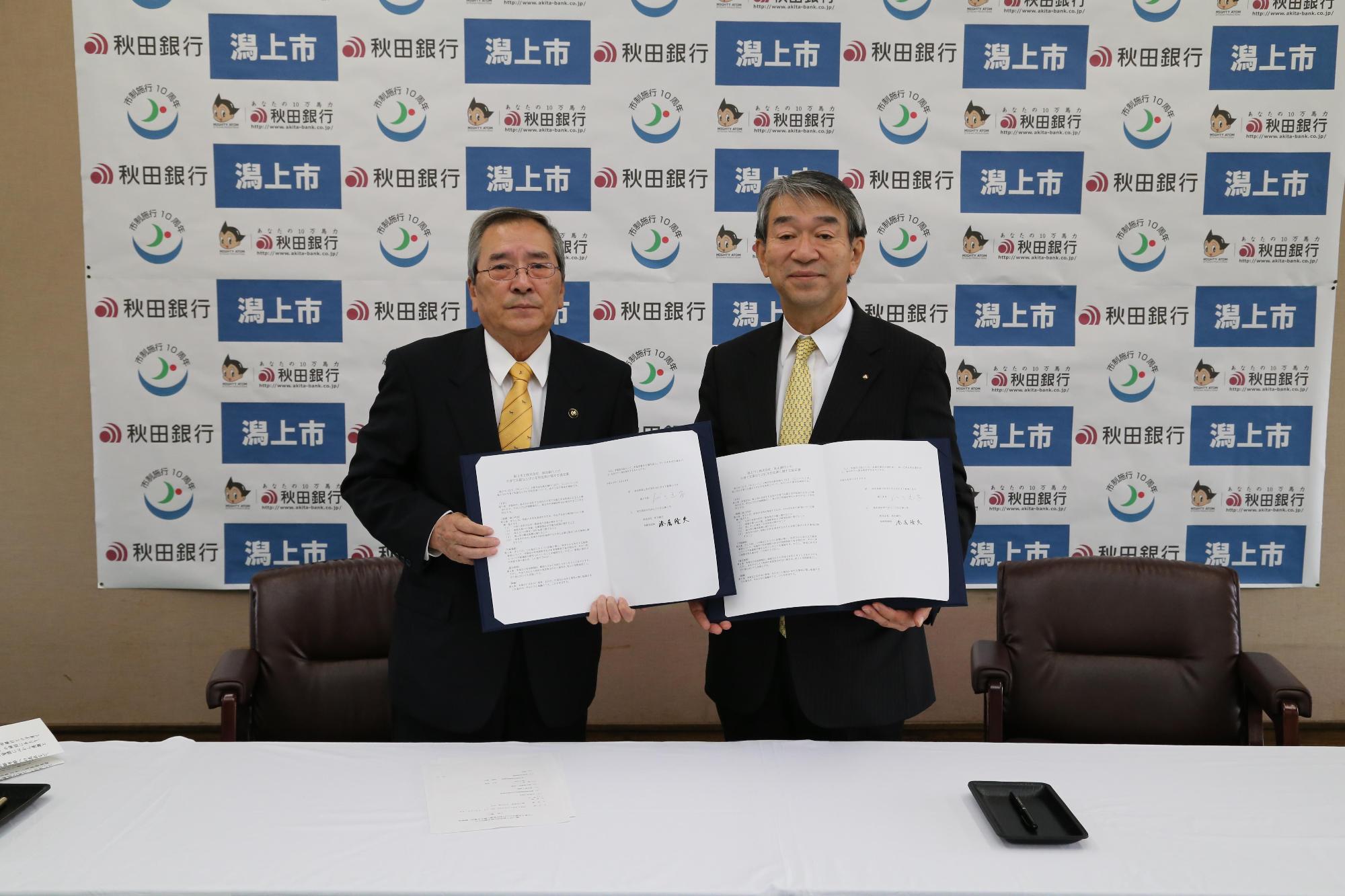 潟上市と秋田銀行と書かれた壁紙の前でスーツを着た二人の男性が両手に協定書を持っている写真